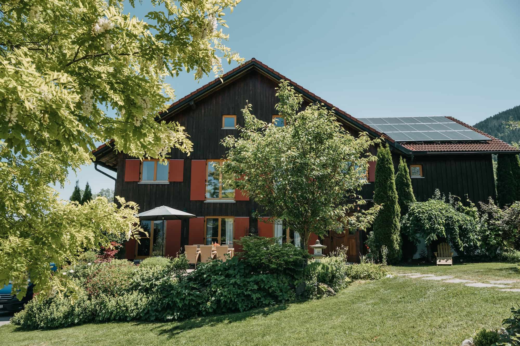 Das dunkle Holzhaus des Mika Hofs steht zwischen Bäumen und Gartenpflanzen.