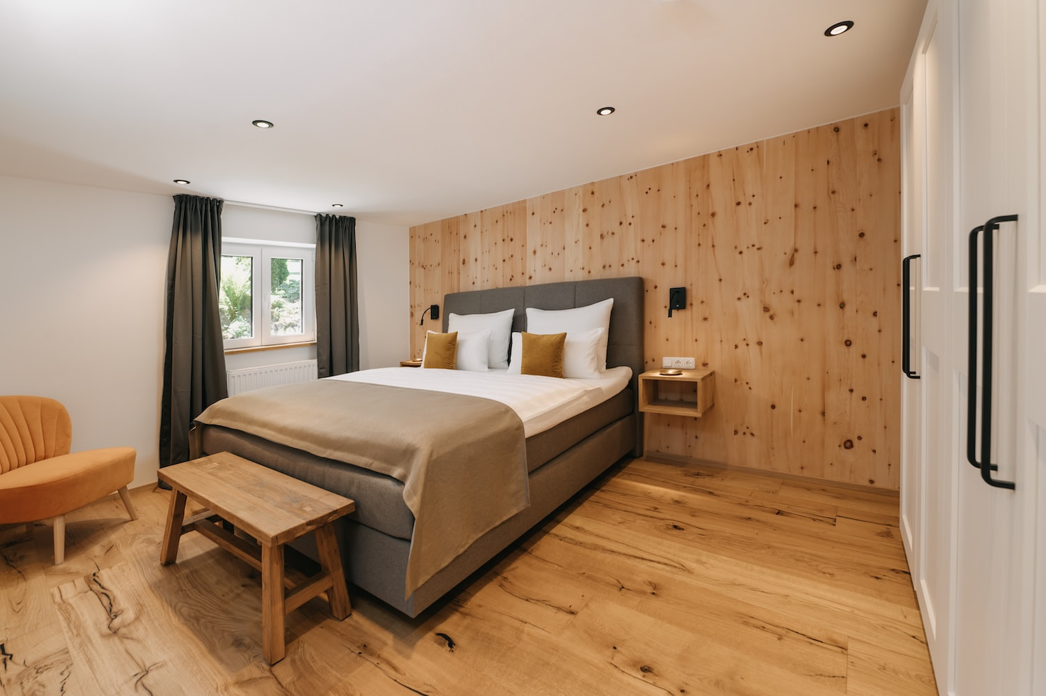 Ein graues Doppelbett steht vor einer Holzwand, daneben ein Fenster mit dunklen Vorhängen.